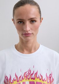 Primo piano di una donna con i capelli raccolti che indossa una camicia bianca con grafiche di fiamme rosa e gialle su uno sfondo semplice.