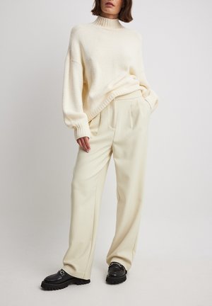 Femme portant un pull col roulé crème, un pantalon ample crème et des mocassins noirs, debout devant un fond clair.