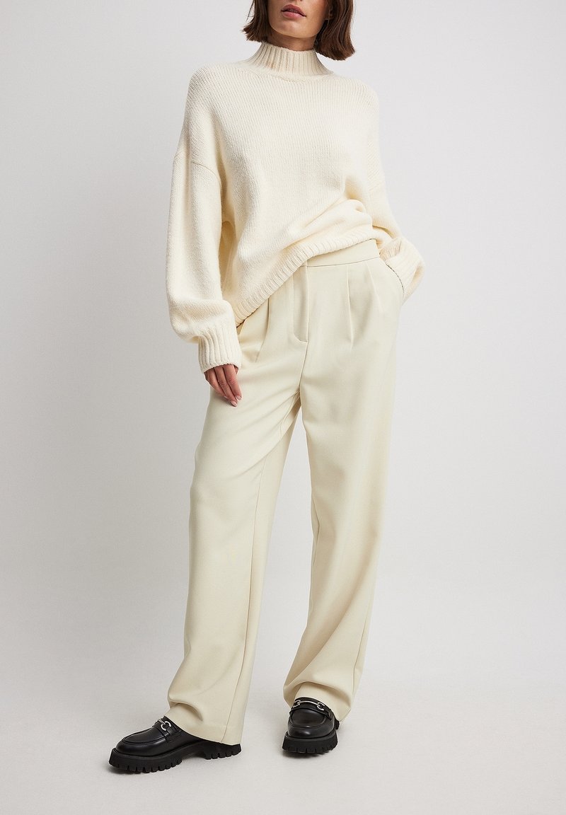 Femme portant un pull col roulé crème, un pantalon ample crème et des mocassins noirs, debout devant un fond clair.