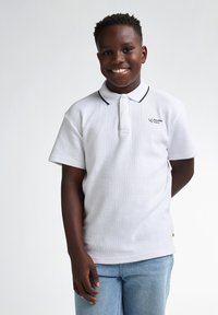 Vit polo med strukturerad tyg, svart krage och en liten logotyp på bröstet. Kombinerad med ljusblå jeans.