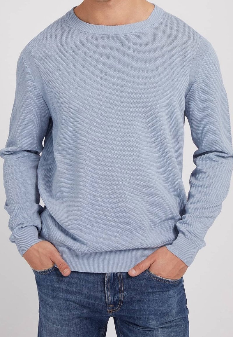 Homme portant un pull à col rond texturé bleu clair et un jean bleu, les mains dans les poches, devant un fond uni.