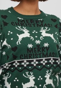 Grön stickad tröja med vita renar, snöflingor och texten "GOD JUL!" med svarta detaljer och rund halsringning.