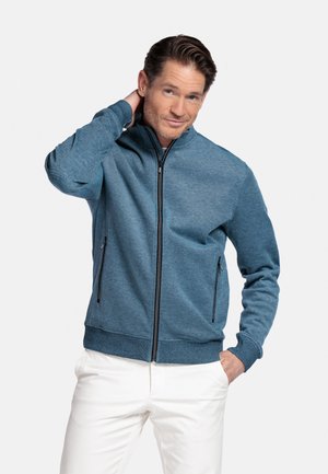 Man in een blauw jack met ritssluiting en zijzakken en witte broek, hand achter de nek, andere hand in de zak, neutrale lichte achtergrond.