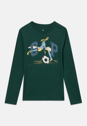 Chemise à manches longues vert foncé avec un graphisme d'un joueur de football donnant un coup de pied dans un ballon entre les grandes lettres G et P.