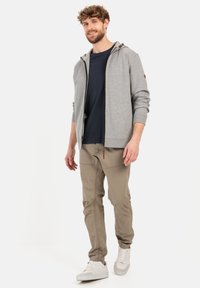 Grijze zip-up hoodie met een capuchon, gecombineerd met een marineblauw shirt en beige broek. Witte sneakers met minimale details maken de outfit af.