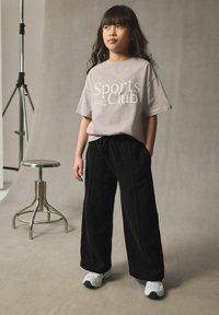 Hellgraues T-Shirt mit dem Aufdruck "Sports Club 1982", kombiniert mit schwarzen weit geschnittenen Hosen. Das Modell trägt weiße Sneaker. Der Hintergrund ist neutral.