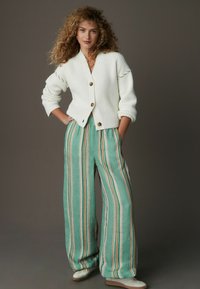 Weißer gerippter Cardigan mit großen Knöpfen, kombiniert mit grün gestreiften, weiten Hosen. Das Outfit zeichnet sich durch weiche Texturen und eine lockere Silhouette aus.