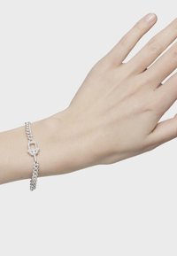 Pulsera de cadena de plata con eslabones entrelazados y un detalle de clasp central, exhibida en una mano sobre un fondo blanco.