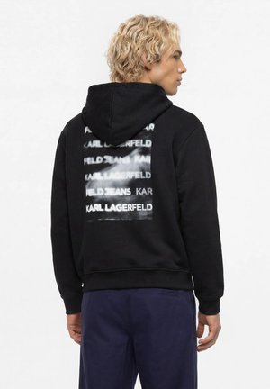 Blonder Mann im Seitenprofil trägt schwarzen Kapuzenpullover mit verschwommenem weißem Textblock auf dem Rücken, der "Karl Lagerfeld" mehrfach zeigt.