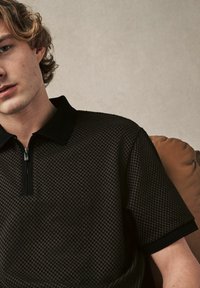 Polo noir avec un motif texturé, col zippé et manches courtes. Le matériau semble léger et respirant.