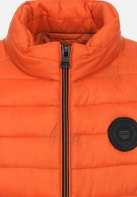 Oranje puffervest met hoge kraag, zwarte rits en zwart, rond logo-patch. Heeft een gewatteerde textuur en een gladde stofafwerking.