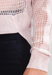 Pull en tricot rose clair avec un motif en mailles ouvertes texturé. Manches longues avec des poignets côtelés et un ourlet légèrement arrondi.