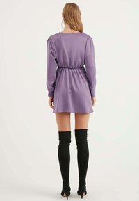 Lila Satin-Minikleid mit langen Ärmeln, elastischer Taille und A-Linie-Rock. Kombiniert mit schwarzen Overknee-Socken und Stiletto-Absätzen.