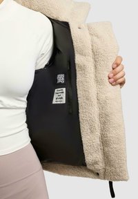 Person trägt ein weißes Oberteil und hält eine beige Fleecejacke offen, die ein schwarzes Innenfutter zeigt, mit Markenetiketten und einer inneren Reißverschlusstasche.