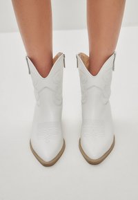 Botas de vaquero blancas con puntas afiladas, patrones de costura intrincados y acabado de piel; cuentan con un cierre de cremallera y suelas en tono marrón.