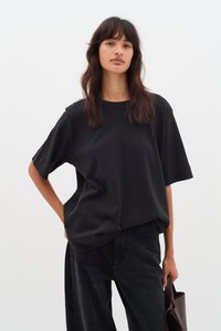 T-shirt nera oversize realizzata in tessuto morbido con maniche corte e colletto rotondo. Indossata con jeans scuri e una borsa marrone.