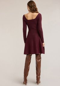 Bordeauxrotes, geripptes Strickkleid mit tailliertem Oberteil und ausgestelltem Rock, ausgestattet mit einem quadratischen Ausschnitt und langen Ärmeln, kombiniert mit braunen Stiefeln, die über das Knie reichen.