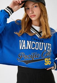 Przycięty niebieski sweter z napisem "Vancouver Pacific Coast 96" w białym i czarnym kolorze, z paskowanym czarnym i białym ściągaczem przy kołnierzyku i mankietach.