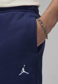 Tumš zilie sporta bikses, izgatavotas no mīksta auduma, ar baltu izšūtu Jumpman logotipu un sānu kabatu ar redzamām šuvju detaļām.