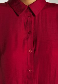 Červená blůza s límečkem z hladké tkaniny, s knoflíkovým zapínáním a odpovídajícími knoflíky. Textura působí lehce a mírně leskle.