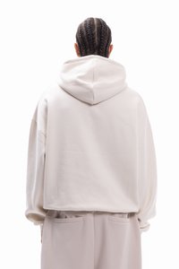 Cremefarbener, kurz geschnittener Hoodie mit großer Kapuze, überschnittenen Schultern und weicher Textur. Zwei seitliche Taschen auf Taillenhöhe sichtbar.