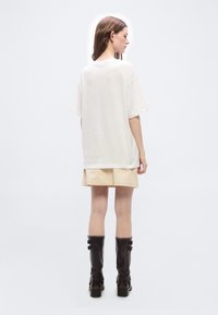 Γυναίκα που στέκεται με την πλάτη προς τα εμπρός, φορώντας μια υπερμεγέθη λευκή μπλούζα, μπεζ σορτς και μαύρες μπότες πάνω από το γόνατο, μπροστά σε απλό φόντο.