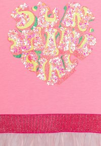 Różowa koszula z błyszczącym wzorem serca zdobionym wielokolorowym brokatem i cekinami, uzupełniona błyszczącym, różowym, prążkowanym dołem.