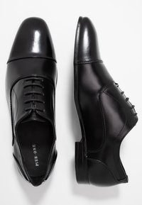 Chaussures habillées en cuir noir avec une finition brillante, dotées d'un bout arrondi, d'un laçage à l'avant et de coutures décoratives sur le côté.