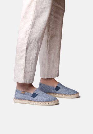 Noge v svetlo modrih espadrilah na natikanje z bež tkanimi podplati in bež posekanimi lanenimi hlačami.