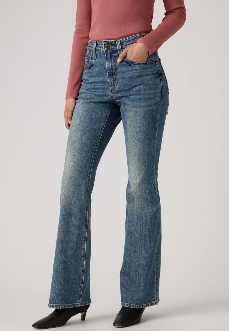 Τζιν με ψηλή μέση και καμπάνα σε ανοιχτό μπλε ντένιμ, με μπροστινές τσέπες, ξεθωριασμένο πλύσιμο και ίσια γραμμή στο παντελόνι. Συνδυασμένο με μαύρες μπότες με τακούνι.