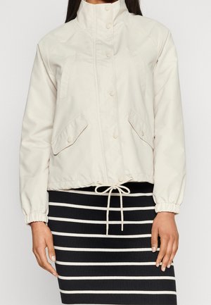 Nainen pukeutuneena kermaan vivahtavaan vetoketjutakkiin, jossa on painonapit, etutaskujen läpät, joustavat hihansuut ja mustavalkoraidallinen hame.