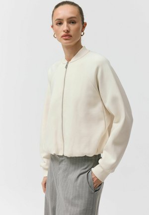 Kobieta ubrana w białą bomberkę na suwak i szare spodnie w prążki, stojąca z jedną ręką w kieszeni na jednolitym tle.