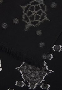 Un material textil negru cu margini franjurate prezintă un model texturat format din forme circulare geometrice, ascuțite, și puncte mici rotunde în nuanțe de alb și gri.