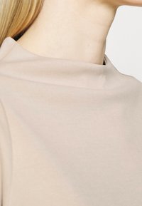 Beżowa bluzka o g�ładkiej teksturze, z wysokim kołnierzem. Materiał jest lekki i miękko opada wokół dekoltu.