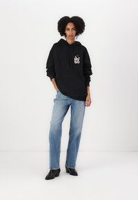 Svart hettegenser med frontlomme og logo; parret med falmede blå jeans og svarte høye støvler, satt mot en hvit bakgrunn.