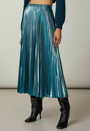 Jupe plissée teal en tissu métallique brillant, avec une taille haute et un design fluide. Associée à des bottes noires à talons.