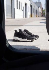 Paire de baskets noires avec des semelles blanches épaisses placées sur un trottoir urbain ensoleillé vues de l'intérieur d'un véhicule.