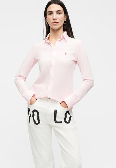 Γυναίκα που φοράει ανοιχτό ροζ πουκάμισο με κουμπιά και λογότυπο στο στήθος και λευκό παντελόνι με μεγάλα μαύρα γράμματα που σχηματίζουν "POLO".