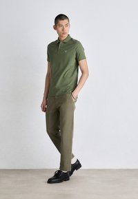 Olíva zöld, rövid ujjú poloing, világosbarna nadrággal és fekete cipővel párosítva. Egyszerű dizájn, egyenes fazon, minimális márkázás.