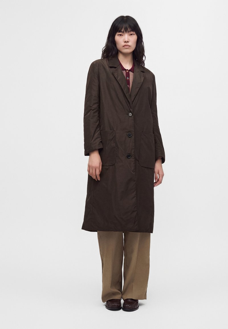 Femme portant un long manteau marron foncé par-dessus un haut bordeaux, un pantalon large beige et des chaussures marron foncé, debout devant un fond uni.