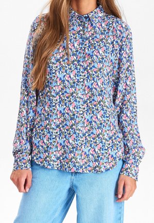 Kvinde iført en langærmet bluse med blomsterprint i blå, pink, hvid og grønne mønstre, kombineret med lyseblå jeans.