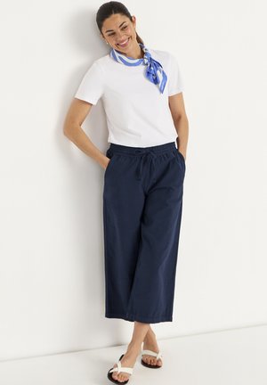 Lachende vrouw die tegen een muur leunt, draagt een wit shirt met korte mouwen, blauwe wijde cropped broek, witte sandalen en een blauw sjaaltje met patroon om haar nek.