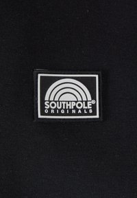 Musta kangas, jossa on suorakulmainen paikka, jossa on valkoinen sateenkaari -kuvio tekstin "SOUTHPOLE® ORIGINALS" yläpuolella paksuilla kirjaimilla.