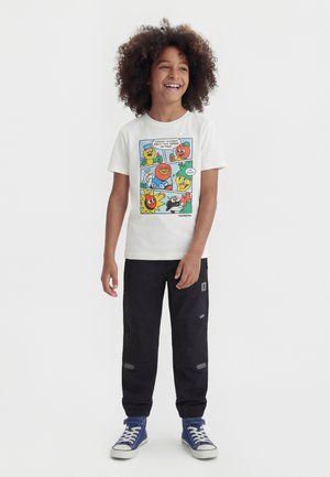 Dziecko o kręconych włosach uśmiechające się, ubrane w białą koszulkę z kolorowymi postaciami owoców i warzyw z kreskówki, czarne spodnie i niebieskie trampki.