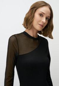 Top nero a maniche lunghe con pannello in rete trasparente su una spalla. Tessuto morbido con silhouette aderente. Design minimalista e colori neutri.