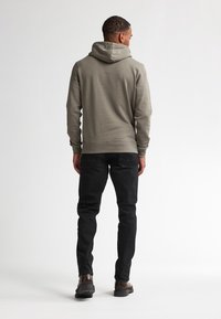Hombre vestido con una sudadera gris, jeans negros y zapatos marrones, de pie de espaldas contra un fondo blanco liso.