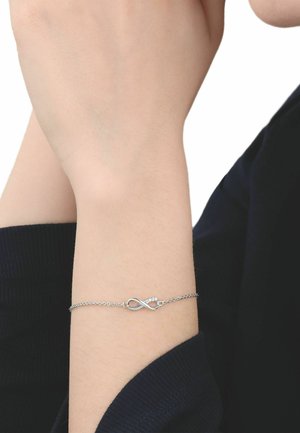 Silbernes Armband mit Unendlichkeitszeichen am Handgelenk, getragen von einer Person mit heller Haut und schwarzem Langarmshirt.