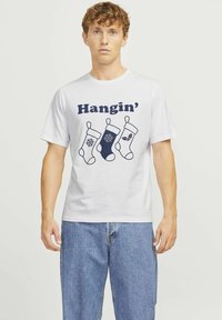 Bijela pamučna majica s tri ilustrirane božićne čarape u plavoj i tamnoplavoj boji, s tekstom "Hangin’" iznad dizajna.