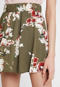 Gonna olive fino al mezzo coscia con vita elasticizzata e grande stampa floreale bianca e rossa, indossata con un top rosa chiaro.
