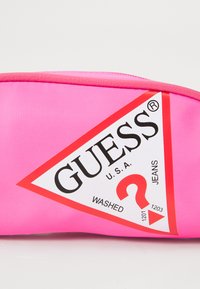 Rózsaszín szövet tasak, háromszög alakú fehér és piros Guess logóval, amelyen a "U.S.A.", "WASHED" és egy stilizált kérdőjel látható.
